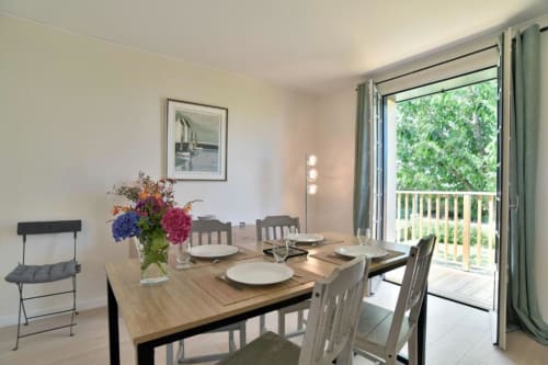 Salle à manger lumineuse de la villa Maison Grand Large avec table en bois, vue sur le jardin par les portes-fenêtres, fleurs fraîches.