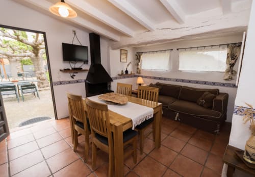 Sala de estar rústica con mesa de comedor de madera, sofá oscuro, chimenea y vista al jardín de la masía en Girona