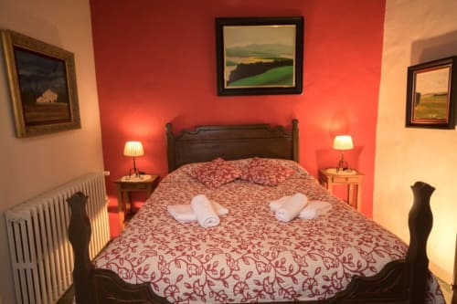 Dormitorio con pared de acento rojo, cama de madera oscura, toallas blancas enrolladas, cuadros campestres, lámparas de mesilla y vistas al jardín