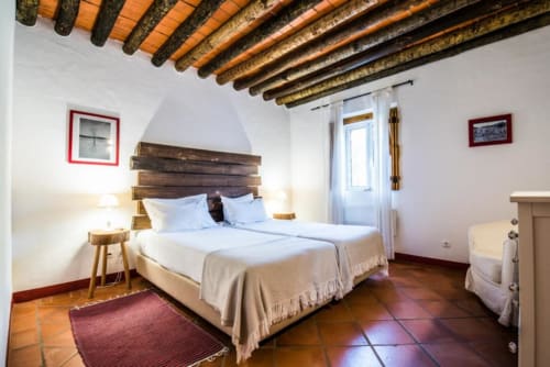 Quarto rústico com teto de vigas de madeira, roupa de cama branca e piso de terracota na villa Casas de Juromenha em Portugal