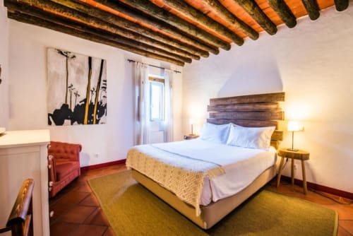 Quarto da villa Casas de Juromenha com vigas de madeira, pisos de terracota, cama, cadeira vermelha e vista para o jardim
