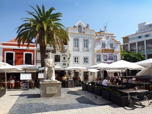 Praça do hotel Cascais Houses em Juromenha com edifícios históricos coloridos, palmeira, estátua e área de estar ao ar livre