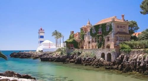 Mansão costeira com paredes cobertas de hera com vista para a baía turquesa, farol e palmeiras em Cascais, Portugal
