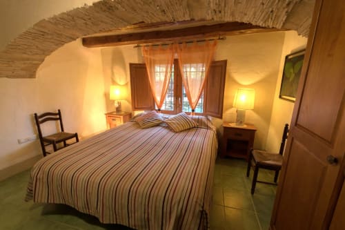 Dormitorio rústico en la masía Can Gat Vell del siglo XVIII en Girona, con ropa de cama a rayas, vigas de madera e iluminación cálida.