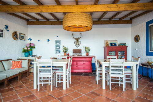 Sala de jantar mediterrânea clara com cadeiras brancas, aparador vermelho e luminária pendente trançada na villa de Juromenha