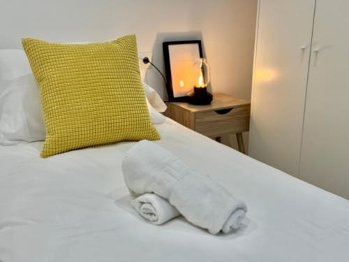 Habitación moderna en Nerja Holiday Homes con almohada amarilla, ropa de cama blanca, toallas enrolladas y mesita de noche con lámpara