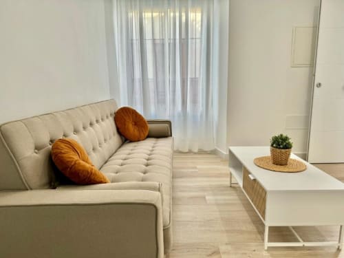 Luminoso salón del apartamento en Nerja con sofá crema, cojines naranjas, planta en maceta y cortinas translúcidas