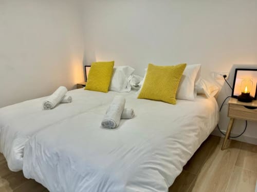 Dormitorio moderno en Nerja con sábanas blancas, cojines amarillos, toallas enrolladas e iluminación ambiental de lámparas mesita de noche