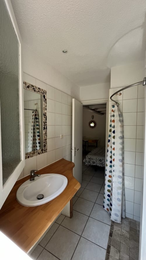 Salle de bain du studio de la villa Le Maquis Plage avec meuble lavabo en bois, rideau de douche à pois et sol carrelé à Poggio-Mezzana
