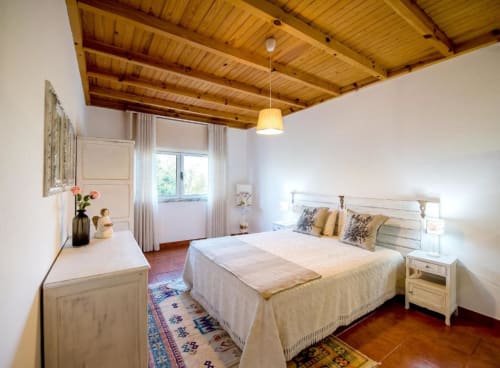 Quarto luminoso na Home Alliance em Bombarral com vigas de madeira no teto, paredes brancas, cama de casal e piso em terracota.