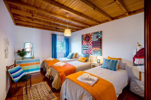 Bright triple bedroom with three beds, wooden ceiling, colorful dresser, blue curtains, and modern decor in Bombarral, Óbidos