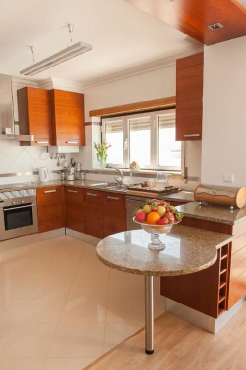 Cozinha moderna com armários em madeira laranja, ilha com bancada em granito e fruteira, eletrodomésticos em aço inox, e vistas para o mar