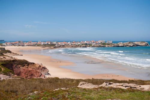 Praia da Baleal com areia dourada, ondas turquesas e vila costeira ao fundo sob céu azul cristalino