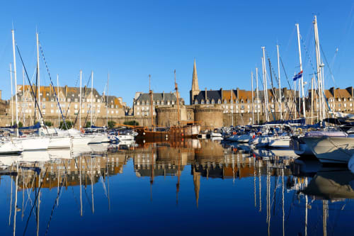 Voiliers amarrés dans le port de Saint-Cast avec château médiéval et bâtiments en pierre reflétés dans l'eau bleue calme