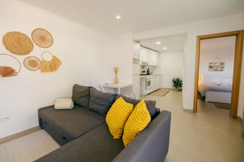 Modern villa living area with gray sofa, yellow cushions, woven wall decor, open-plan kitchen, and bedroom at Tree House