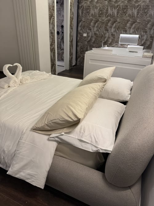 Camera moderna presso Enjoy House Bracciano con cuscini bianchi, letto con testata imbottita grigia, cigni di asciugamani e zona living