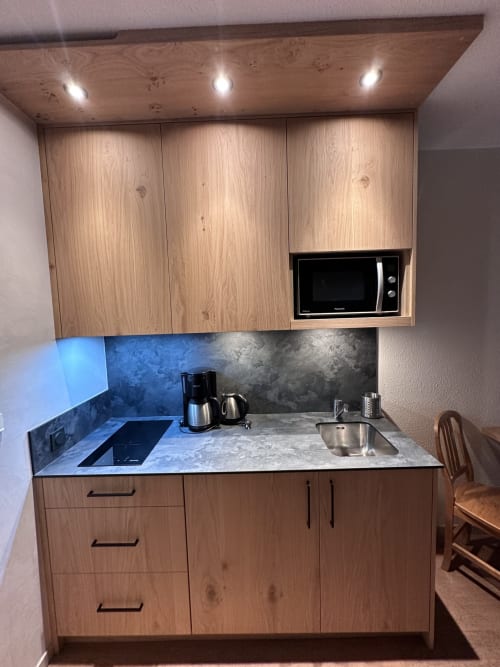 Kitchenette moderne avec mobilier en bois clair, plan de travail gris, micro-ondes et dosseret bleu à Hôtel Studios La