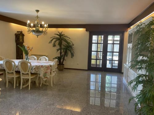 Espaçosa sala de jantar da Casa Da Avó Ilda com toalha de mesa floral, cadeiras creme, lustre, plantas em vasos e portas de vidro