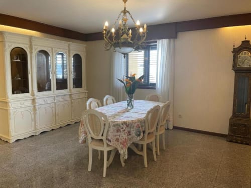 Elegante sala de jantar da Casa Da Avó Ilda com mesa redonda, cadeiras creme, lustre de cristal e cristaleira branca.