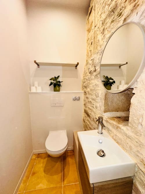 Salle de bain moderne à Vue Sur Gindou avec lavabo blanc, miroir rond, mur en pierre, toilettes et carrelage doré