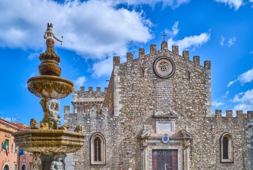 Chiesa medievale in pietra con merli e torre orologio, fontana decorativa in primo piano, Taormina
