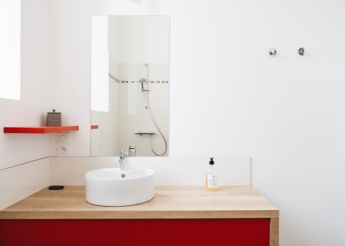 Salle de bain moderne du gîte La Maison D'olivier avec lavabo rond blanc, meuble en bois, étagère rouge et douche à l'italienne vitrée