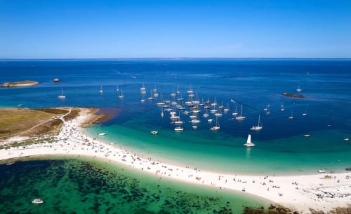 Aerial view of Concarneau's turquoise bay with white sand beach, moored sailboats, and rocky islands in Brittany