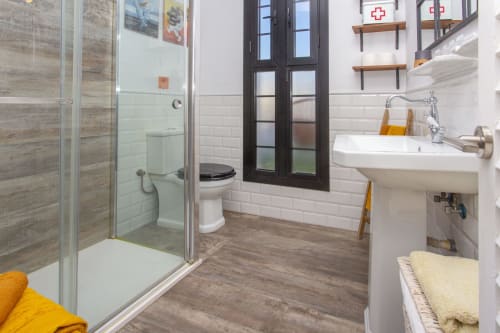Baño moderno con cabina de ducha de cristal, lavamanos blanco, inodoro, suelo de madera y estantería del apartamento Wehost Costaluz