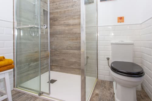 Baño moderno en apartamento Wehost Costaluz, Puerto Santa María, con ducha a ras de suelo, asiento de inodoro negro y azulejos blancos