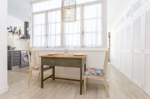 Cocina-comedor luminoso con escritorio verde, sillas de madera, lavadora y grandes ventanas en Puerto Santa María