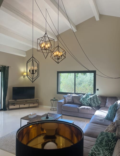 Modernes Villen-Wohnzimmer mit geometrischen Hängeleuchten, grauem Sofa, Kamin und Gartenblick in La Conciergerie De Franck