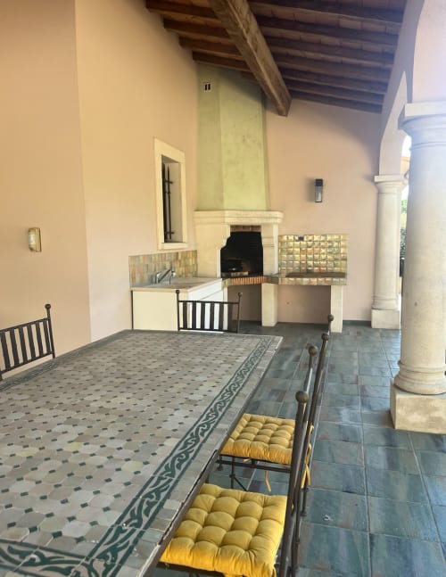Überdachte Terrasse mit gemustertem Fliesenboden, gelben gepolsterten Stühlen, Waschbecken, Kamin und weißen Säulen in La Conciergerie De