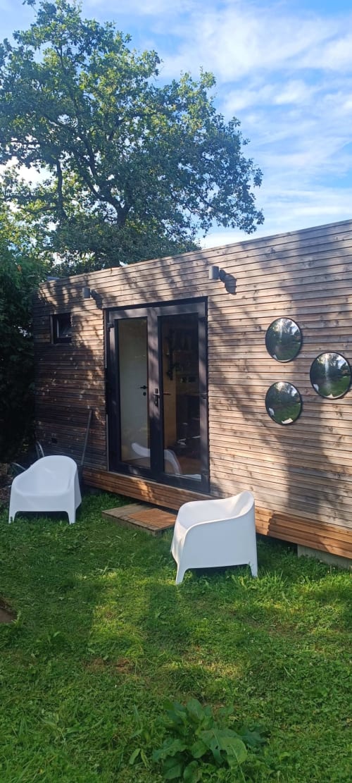 Modern wooden cottage with glass doors and circular windows, white chairs on grass, Jalhay Belgium