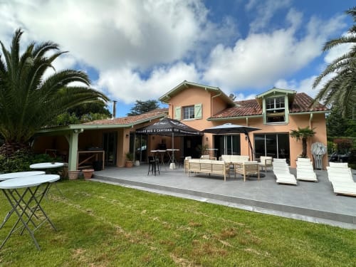 La Conciergerie Biarritz B&B terrace with loungers, black umbrella, palm trees, and peach-colored building.