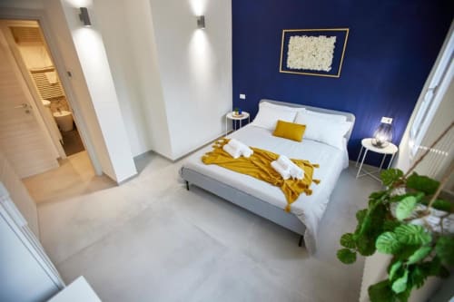 Modern bedroom at L.H.A. hotel in Ferrara with blue accent wall, white bed, gold throw, and ensuite bathroom.