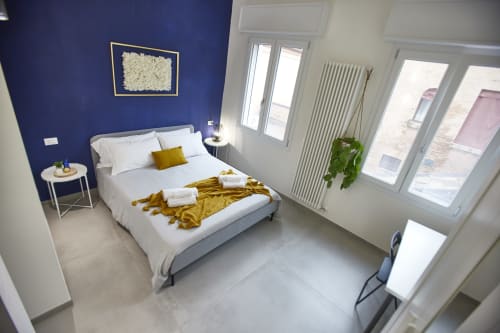 Modern bedroom at LikeHome San Romano in Ferrara with blue accent wall, queen bed, gold throw, city view, and contemporary