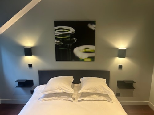Modern bedroom at Le Petit Château with white bedding, dark headboard, wall sconces, and garden view artwork above bed