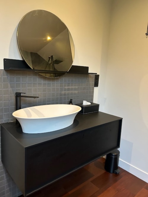 Modern bathroom vanity with white vessel sink, black counter, round mirror, and gray tile backsplash at Le Petit Château in
