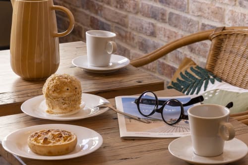 Ambiance chaleureuse du café de l'Hôtel De Lion Sur Mer avec pâtisseries, tasse de café, théière et lunettes de lecture sur table en bois