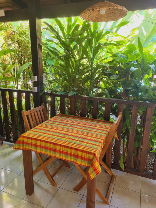 Outdoor dining area at Gites Kaladja with wooden table, colorful plaid tablecloth, chair, woven lamp, lush tropical plants,