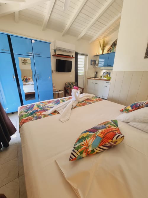 Bright studio bedroom with two single beds, colorful runners, blue wardrobe, kitchenette, and TV at Gites Kaladja Port-Louis