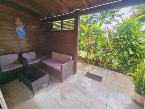 Covered patio with pink wicker seating, tropical plants, and shuttered windows at Gites Kaladja apartment in Port-Louis