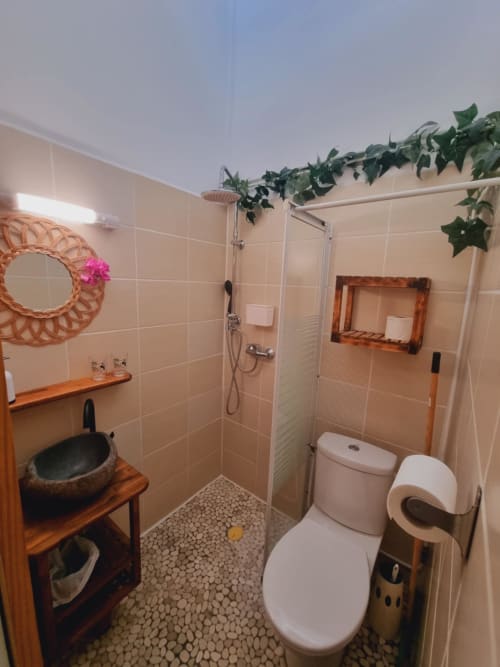 Modern bathroom with walk-in shower, toilet, round black sink on wooden vanity, decorative plants and woven wreath at Gites