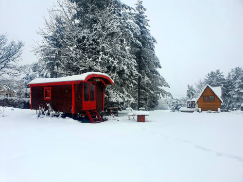 Red caravan in snowy garden at Les Refuges Du Chalet, Jalhay, with pine trees and wooden cabin