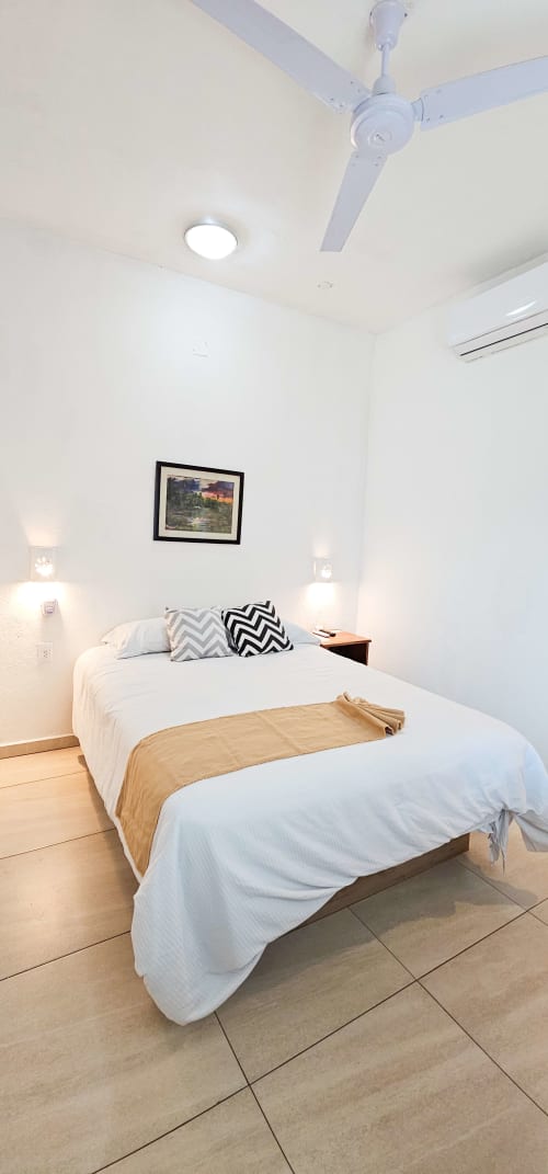 Clean modern bedroom at La Familia Hotel in San Blas, Nayarit, featuring queen bed with white linens, ceiling fan, and wall