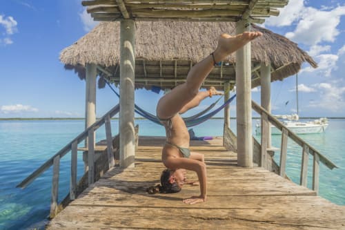 Gimnasta haciendo parada de cabeza en el muelle de madera del hostal Cayuco Maya en Bacalar con agua turquesa