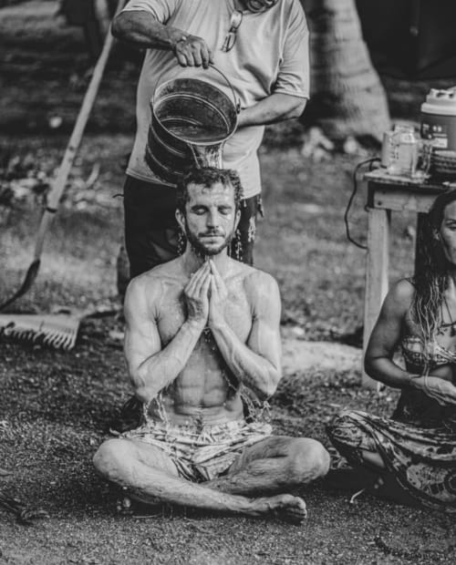 Hombre sin camisa en posición de meditación recibe ducha con cubeta de agua en el hostal Cayuco Maya, Bacalar, México