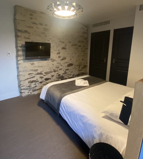 Chambre moderne de l'Hôtel Saint Roch à Saint Ybard avec deux lits, mur en pierre, TV écran plat et portes foncées