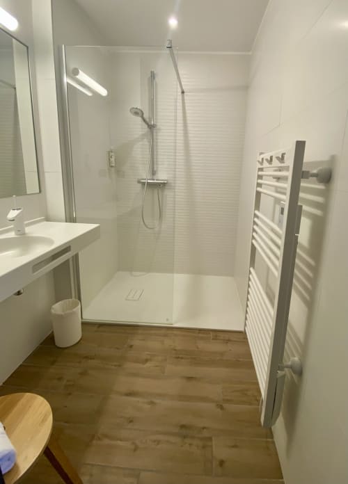 Salle de bain moderne de l'Hôtel Saint Roch avec douche ouverte, vasque blanche, sèche-serviettes chauffant et parquet