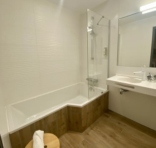 Salle de bain moderne de l'Hôtel Saint Roch avec carrelage blanc métro, baignoire avec cabine de douche vitrée et meuble vasque en bois.